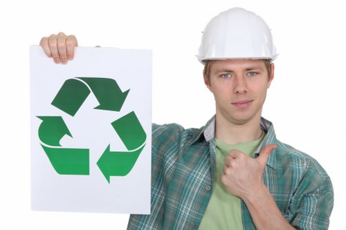 A man wearing a white safety helmet and casual checkered shirt stands outdoors against a plain backdrop, holding a white sign displaying a large green recycling symbol composed of three arrows forming a continuous loop. The man is giving a thumbs-up with his right hand and has a neutral facial expression, suggesting approval or endorsement of recycling practices. The image context implies a focus on waste management and environmentally conscious rubbish removal services, with the recycling symbol highlighting the importance of proper waste disposal. The setting and attire indicate a professional yet approachable environment, appropriate for a commercial waste removal company operating around Epping, though the background remains minimal to keep full attention on the individual and the sign. The overall tone remains neutral and factual, suitable for a website related to rubbish management, emphasizing environmentally responsible waste practices without promotional language.