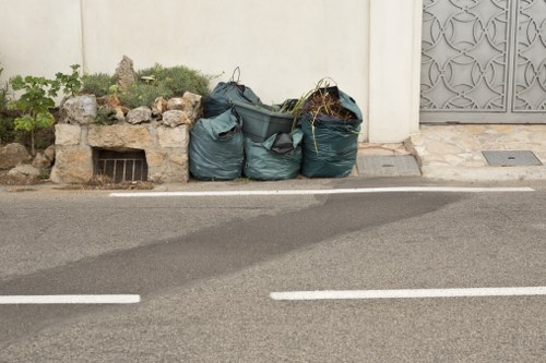 A row of four large, dark green refuse bags filled with waste material positioned on the pavement next to a low stone wall with greenery and rocks behind it. The bags appear to be made of durable plastic, some with visible creases and slight gleam from the light. They are situated close to a white wall and an ornate metal gate on the right side of the image, indicating an urban or residential setting. The bags are aligned along a typical tarmac road with white dashed lines marking the vehicle lanes in the foreground. The surrounding environment includes a small vent or drainage opening beneath the stone wall and minimal vegetation, suggesting a manageable rubbish collection site on a street that might be serviced by local waste removal providers such as Commercial Waste Epping, supporting professional rubbish disposal in the Epping area. The overall scene is well lit, with natural daylight highlighting the textures and colors of the waste bags and urban elements, reflecting a typical rubbish collection or disposal scene in the vicinity of Epping, Essex.