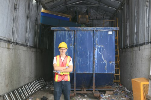 A construction or waste management site inside a large, partially enclosed industrial structure with metal walls and a curved ceiling. In the foreground, a worker wearing a yellow safety helmet, a high-visibility orange and yellow safety vest, and dark work trousers stands with arms crossed, facing the camera. Behind the worker, a large blue metal skip or waste container is positioned centrally, filled with debris and waste materials. The container's surface has visible scratches and weathering. To the right, a yellow plastic waste bin with a slightly textured surface is placed on the concrete floor, which is scattered with small bits of rubbish and paper. On the left side, a black and silver ladder leans against the wall, and on the right, a yellow safety ladder or scaffolding is partly visible next to the container. The environment appears well-lit, with natural light filtering into the space, characteristic of a waste collection or disposal area in Epping, managed by Commercial Waste Epping, with a focus on rubbish collection and disposal activities in the local area.