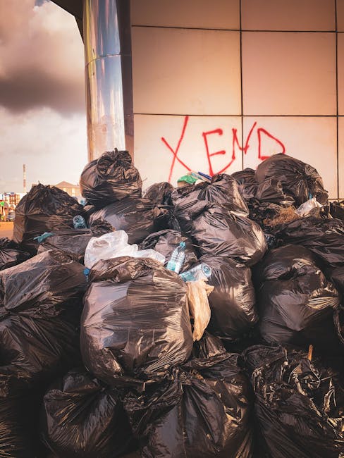 A narrow urban alleyway filled with overflowing rubbish and discarded waste materials, including large black and grey refuse bags, some torn open revealing contents, and a variety of cardboard boxes s