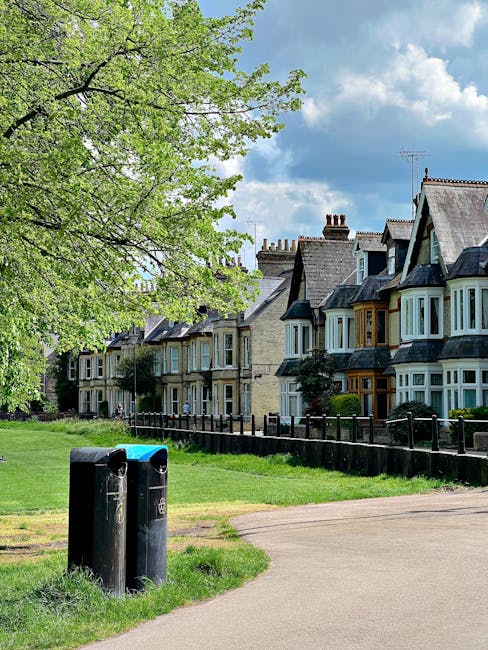 A quiet residential street scene featuring a row of traditional Victorian-style terraced houses with bay windows, pitched tiled roofs, and brick facades, situated behind a front garden enclosed by a l